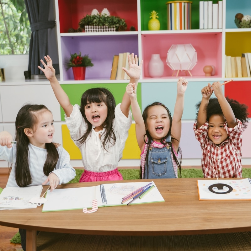 Happy children raising hands while sitting at a table in a colourful daycare centre, enjoying educational activities.