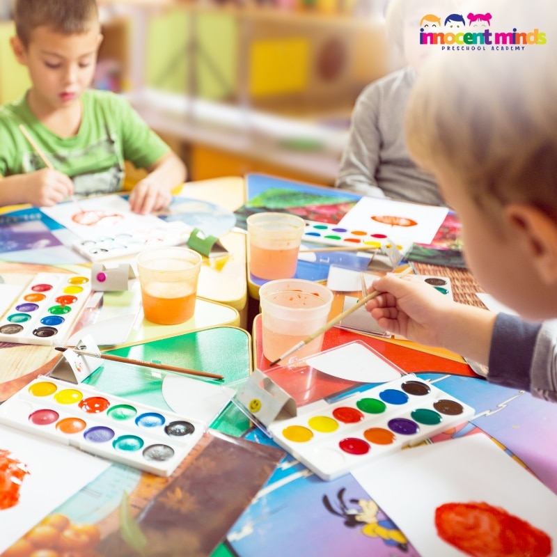 Children engaged in painting at a table, using brushes and vibrant colors in a preschool program setting.