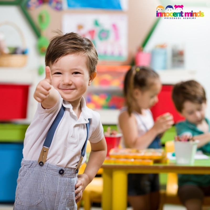 A young boy in a classroom gives a thumbs up, showcasing enthusiasm for learning in a structured preschool environment.