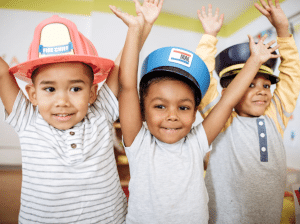 Three young children, who appear to be boys, are smiling and raising their hands in the air. The child on the left is wearing a pink fire chief helmet, the child in the center is wearing a blue mail carrier hat, and the child on the right is wearing a dark blue police officer hat. They seem to be engaged in imaginative play. This could represent an aspect of child care safety through supervised and engaging activities.
